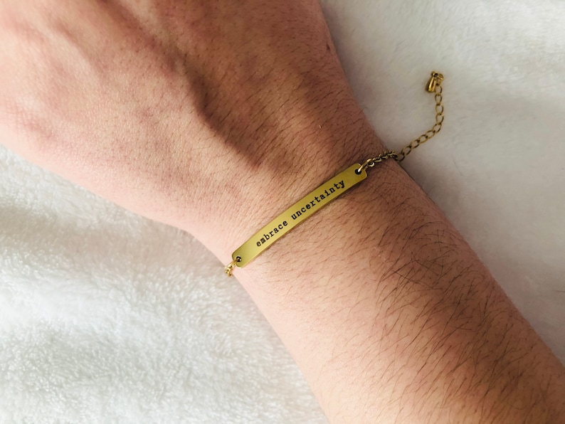 May include: A gold bracelet with a thin chain and a rectangular charm that reads "embrace uncertainty".
