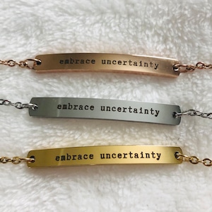 May include: Three silver bar necklaces with the words "embrace uncertainty" engraved on each. The necklaces are on a white fuzzy surface.