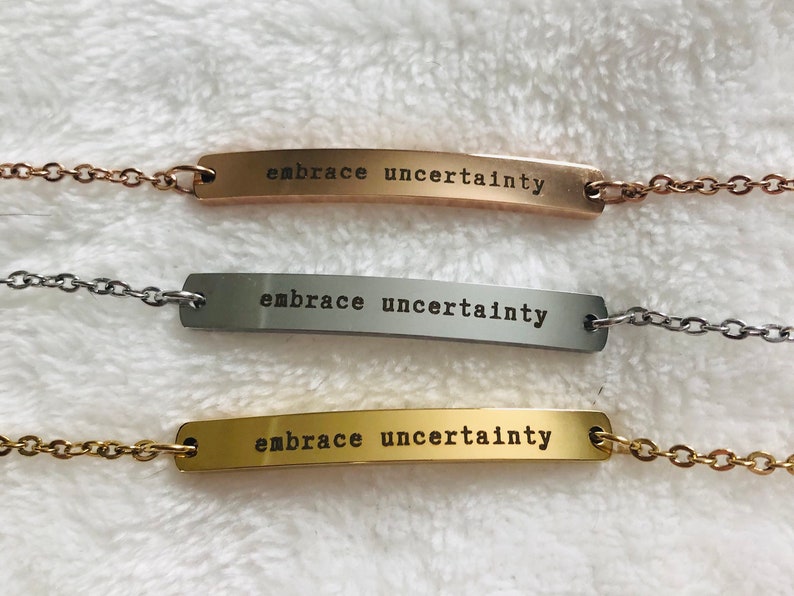 May include: Three silver bar necklaces with the words "embrace uncertainty" engraved on each. The necklaces are on a white fuzzy surface.