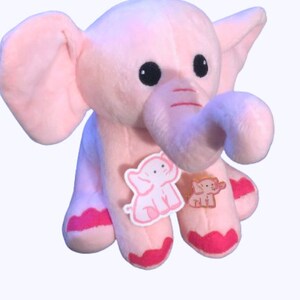 May include: Pink plush elephant toy with a curled trunk and pink accents. The elephant has embroidered eyes and two small elephant appliques on its chest.