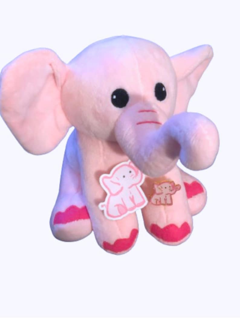 May include: Pink plush elephant toy with a curled trunk and pink accents. The elephant has embroidered eyes and two small elephant appliques on its chest.