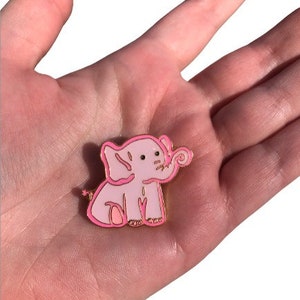 May include: A pink enamel pin featuring a cartoon elephant with a curled trunk. The elephant has a gold outline and a small star on its tail.
