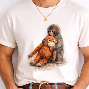 May include: White t-shirt featuring a detailed illustration of a gray monkey hugging an orange stuffed monkey. The artwork is centered on the shirt, showcasing the monkeys in a tender embrace. The shirt is made of a soft material.