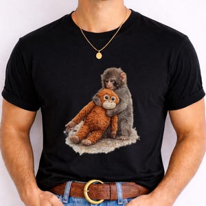 May include: Black t-shirt featuring a detailed illustration of a gray monkey hugging an orange stuffed monkey. The design is printed on a black t-shirt, creating a striking contrast. The image is centered on the shirt.