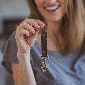 May include: A brown leather key chain with a brass ring and a brass clip. The key chain is held in a person's hand.