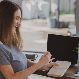 May include: A woman smiles while opening a brown gift box tied with twine. The box is sitting on a wooden table next to a laptop and a cup of coffee.