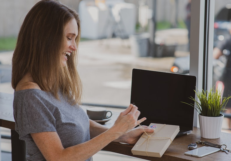 May include: A woman smiles while opening a brown gift box tied with twine. The box is sitting on a wooden table next to a laptop and a cup of coffee.