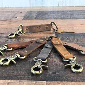 May include: Six leather keychains with brass clasps and key rings. The keychains are in various shades of brown and gray. One keychain has the name "SAK" engraved on it.