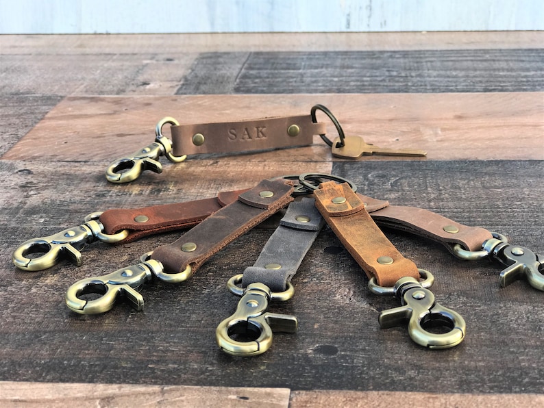 May include: Six leather keychains with brass clasps and key rings. The keychains are in various shades of brown and gray. One keychain has the name "SAK" engraved on it.