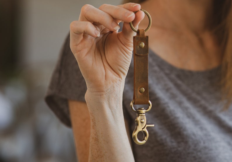 May include: A brown leather keychain with a brass key ring and a brass hook. The keychain is held in a person's hand.