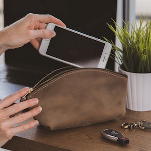 May include: A brown leather zippered pouch with a gold zipper. A hand is placing a white smartphone into the pouch. A white potted plant with green leaves is in the background.