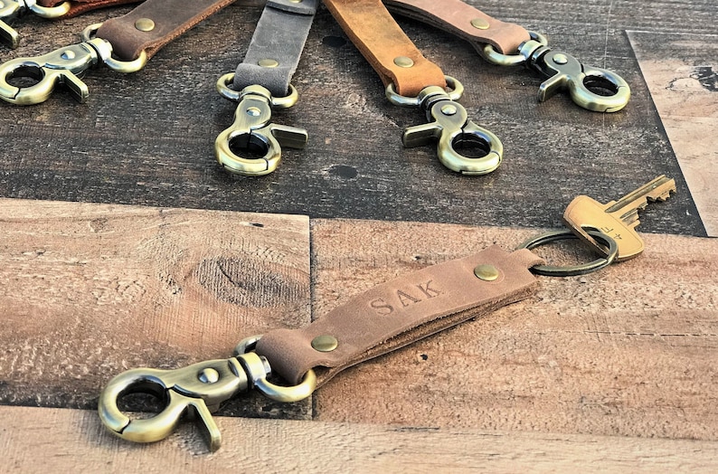 May include: A set of six leather keychains with antique brass clasps and key rings. One keychain has the name "SAK" embossed on the leather.