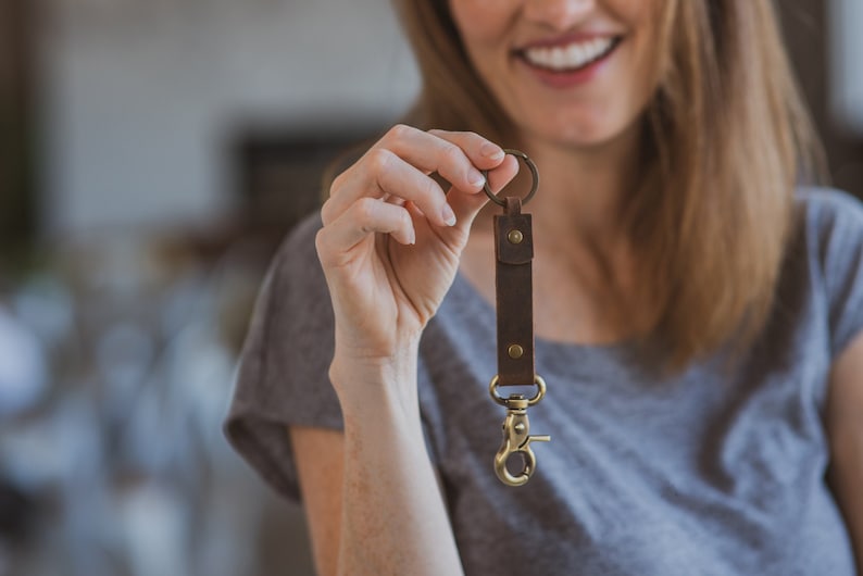 May include: A brown leather key chain with a brass ring and a brass clip. The key chain is held in a person's hand.