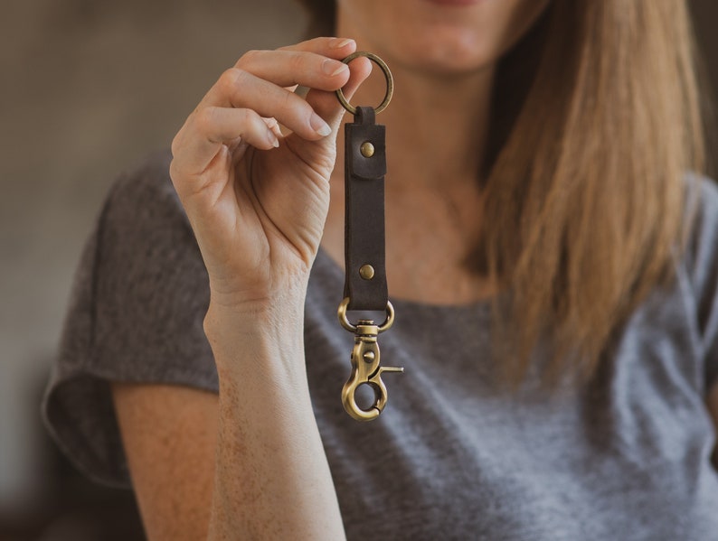 May include: A brown leather key chain with a gold metal ring and a gold metal clip. The key chain is held in a person's hand.