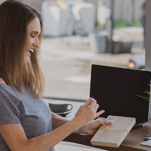 May include: A woman smiles while opening a brown gift box tied with twine. The box is sitting on a wooden table next to a laptop and a cup of coffee.