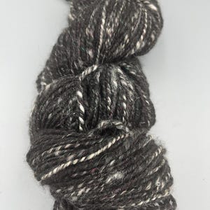 May include: A skein of black yarn with white and pink flecks. The yarn is made of wool and is suitable for knitting or crocheting.