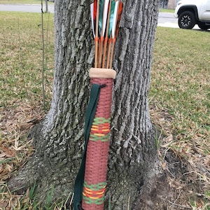 May include: A woven brown quiver with a green strap, holding several arrows with orange, green, and white fletching. The quiver is leaning against a tree trunk, showcasing a rustic, handcrafted design.