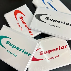 May include: Four white stamp pads with the word "Superior" and "Stamp Pad" printed on them. The stamp pads are in different colors: red, black, blue, and green.
