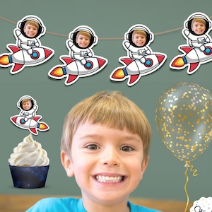 May include: A birthday party banner with a rocket ship and astronaut theme. The banner features a child's face in the astronaut's helmet. The banner is strung on a white string. There is a cupcake with a rocket ship and astronaut topper. There is a gold confetti balloon.