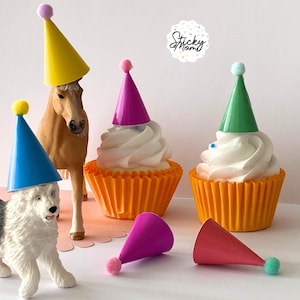 Puede incluir: Una colección de sombreros de fiesta en miniatura amarillos, azules, morados, verdes y rosas, cada uno con un pequeño pompón. Los sombreros se muestran sobre una figura de perro, una de caballo y dos cupcakes en moldes naranjas.