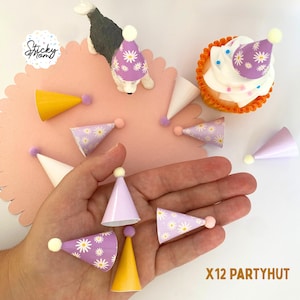 Può includere: Un set di dodici cappellini da festa in miniatura di vari colori e fantasie, tra cui viola con margherite bianche, giallo tinta unita e bianco tinta unita. Ogni cappello conico è sormontato da un piccolo pon-pon. Il testo X12 PARTYHAT è visibile.