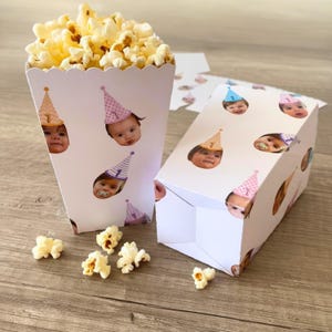 Può includere: Un contenitore e una scatola di popcorn bianchi decorati con facce di bambini con cappellini da festa. Il contenitore per popcorn è pieno di popcorn. La scatola e il contenitore sono su una superficie di legno.