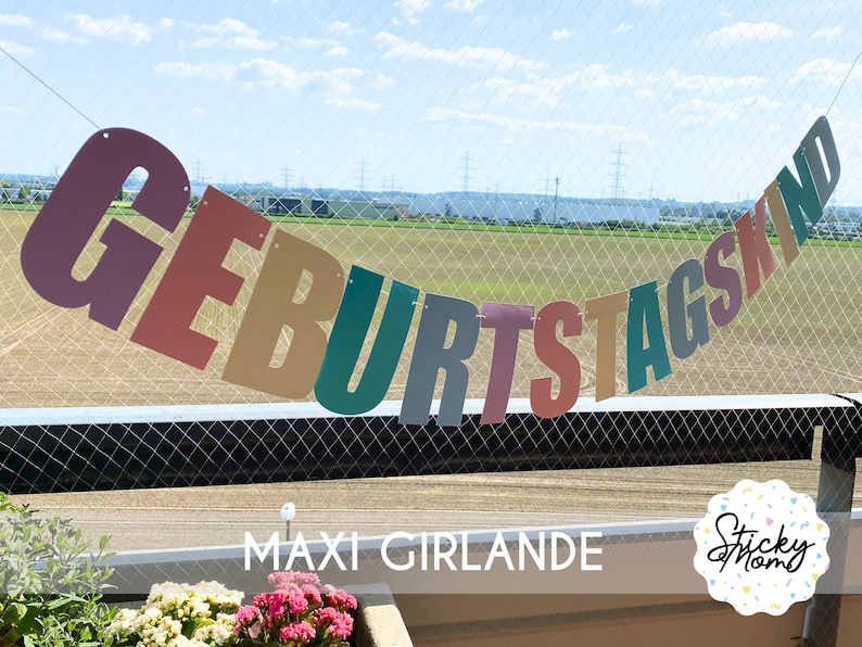 May include: A colourful banner with the text "GEBURTSTAGSKIND" hanging against a backdrop of a green field and a blue sky. The banner is made of individual letters, each a different colour. The text "MAXI GIRLANDE" is printed below the banner.