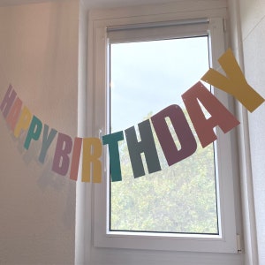 May include: A colourful Happy Birthday banner with letters in different colours, including yellow, green, pink, and purple, hanging in front of a window.