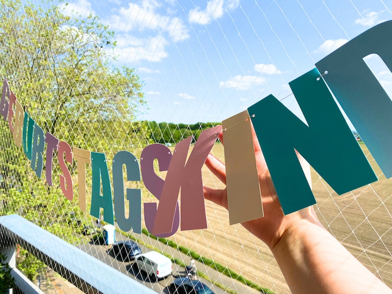 May include: A colourful paper banner with the words "Geburtstagskind" strung across a white net. The banner is made of individual letters in different colours, including pink, blue, green, and yellow.
