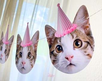 May include: A garland of cat faces, each wearing a pink and white striped party hat. The cat faces are brown, white, and tan, with large, expressive eyes. The garland is strung on a thin white string, perfect for a cat-themed birthday party.