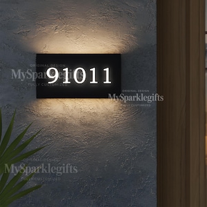 May include: Black rectangular address sign with illuminated white numbers 91011. The sign is mounted on a textured gray wall. The words "ORIGINAL DESIGN" and "MySparklegifts FULLY CUSTOMIZED" are printed on the sign.