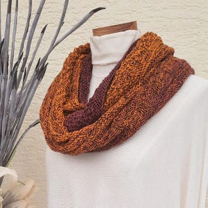 Könnte beinhalten: Ein brauner und orangefarbener gestrickter Infinity-Schal. Der Schal ist aus einem weichen, strukturierten Garn gefertigt und perfekt, um sich in den kälteren Monaten warm zu halten.