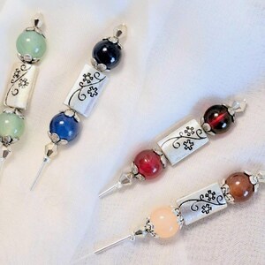 Könnte beinhalten: Vier dekorative Perlenstifte mit silbernen Metallakzenten. Jeder Stift hat ein silbernes Rechteck mit einem floralen Design und eine andersfarbige Perle an jedem Ende. Die Perlen sind grün, blau, rot und braun.