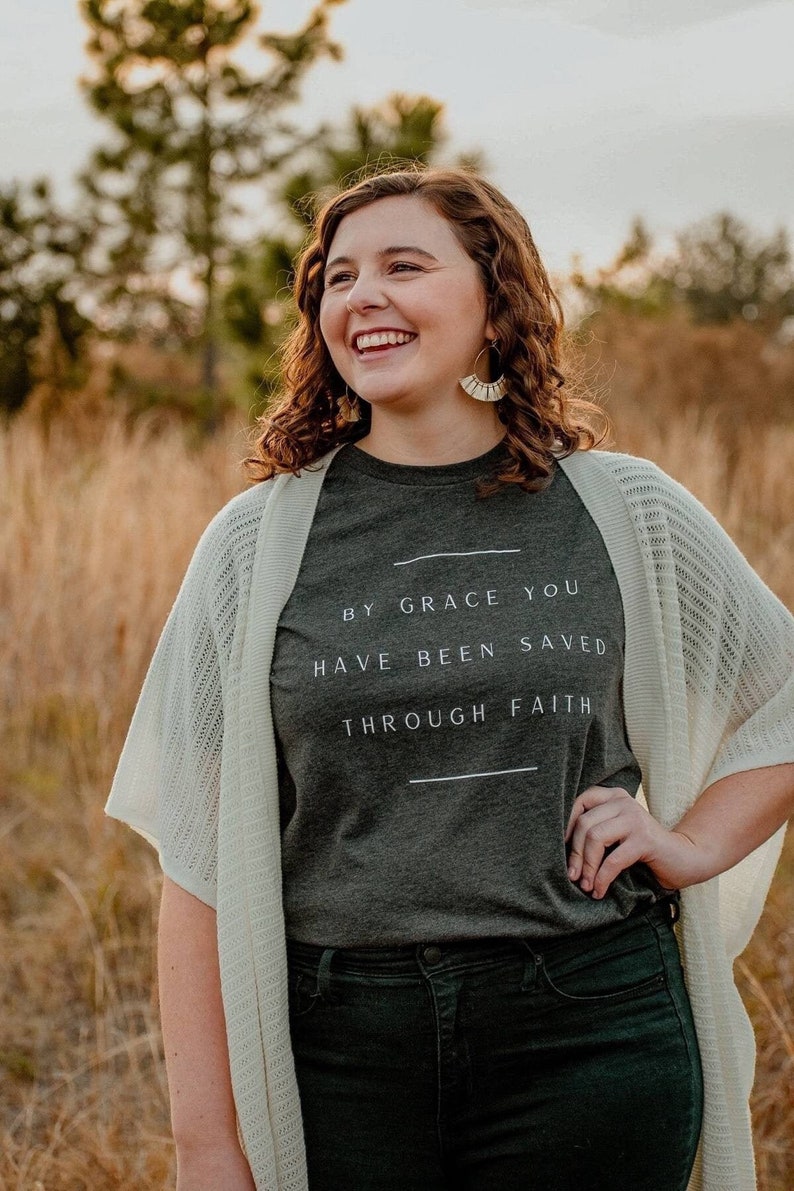 K&ouml;nnte beinhalten: Eine Frau tr&auml;gt ein dunkelgraues T-Shirt mit dem Text "By grace you have been saved through faith" in wei&szlig;en Buchstaben. Sie tr&auml;gt auch einen wei&szlig;en Cardigan und eine dunkelgr&uuml;ne Jeans.