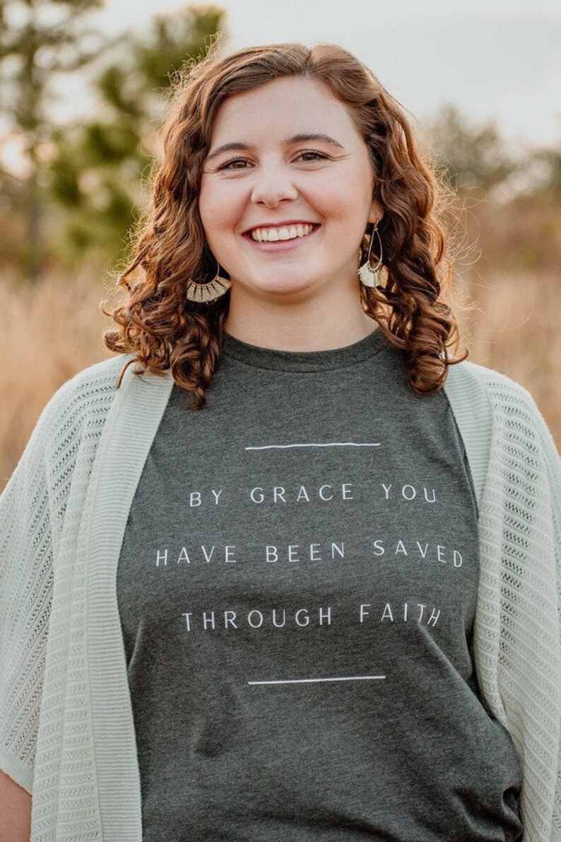 K&ouml;nnte beinhalten: Eine Frau tr&auml;gt ein dunkelgraues T-Shirt mit dem Text "By grace you have been saved through faith" in wei&szlig;en Buchstaben. Sie tr&auml;gt eine hellbeige Strickjacke und hat lockiges braunes Haar.