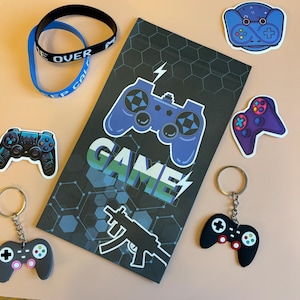 Op de afbeelding: Een zwart notitieboek met een blauwe en groene "GAME"-afbeelding en een blauw ontwerp van een videogamecontroller. Het notitieboek is omringd door verschillende gaming-stickers, waaronder een blauwe en witte controller, een zwarte en groene controller en een zwarte en rode controller-sleutelhanger.