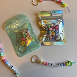May include: A selection of keychains featuring colourful beads in star and flower shapes, spelling out names such as "ISLA", "SOPHIE", and "MOLLY". Some are packaged in iridescent pouches, with extra beads and key rings.