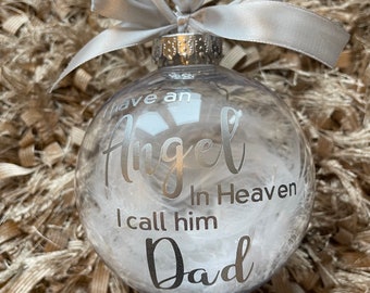 May include: A clear glass Christmas ornament with white feathers inside. The ornament has a silver inscription that reads "I have an Angel in Heaven I call him Dad."