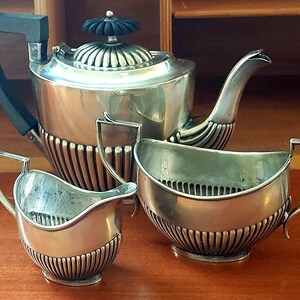 May include: A silver tea set comprising a teapot, creamer and sugar bowl. The teapot features a black knob on the lid and a fluted design. The creamer and sugar bowl also have fluted designs.