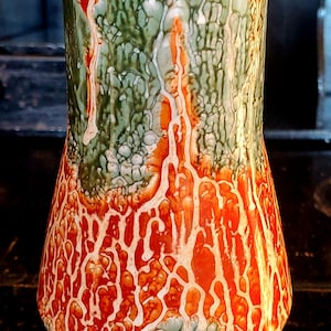 May include: A ceramic vase with a green and orange marbled glaze. The vase has a wide base and a narrow neck.