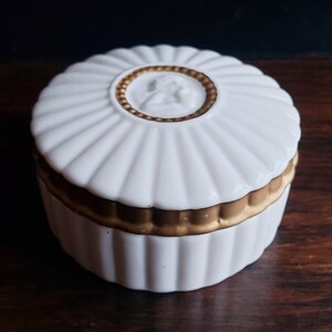 May include: A white ceramic trinket box with a fluted design and a gold-coloured band around the middle. The lid features a gold-coloured border and a central relief of a classical figure. The box is on a dark wooden surface.