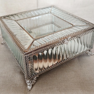 May include: A rectangular mirrored glass jewellery box featuring bevelled edges and a silver-toned metal frame with intricate filigree detailing along the borders. The box stands on four ornate, decorative metal feet and has a hinged lid.