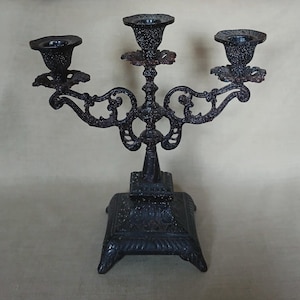 May include: A black metal candelabra with a speckled finish, featuring three candle holders, ornate scrollwork arms, and a square, tiered base with decorative relief patterns. The structure is symmetrical with a central pillar and two side arms.