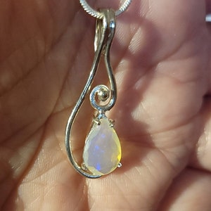 May include: A silver pendant necklace featuring a pear-shaped opal gemstone. The opal displays a play of color with flashes of blue, yellow, and white. The pendant is suspended from a delicate silver chain, showcasing a minimalist design.