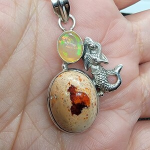 May include: A silver pendant necklace featuring two oval opals and a mermaid charm. The larger opal is beige with orange and brown inclusions. The smaller opal is yellow with green and orange flashes. The mermaid charm is detailed.