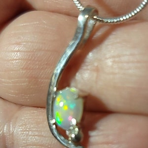 May include: A silver pendant necklace featuring a pear-shaped opal gemstone. The opal displays a play of color with flashes of green, yellow, and pink. The pendant has a curved, flowing design and is suspended from a delicate silver chain.