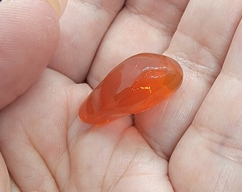 May include: A polished, teardrop-shaped orange gemstone. The translucent stone has a smooth, glossy surface and is held in an open hand. The gemstone's color ranges from light to deep orange, with a slight internal shimmer.
