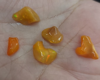 May include: A collection of six polished, orange-yellow opal gemstones. The opals have a smooth, glossy surface and varying shapes, with some displaying flashes of green and yellow iridescence. The gemstones are displayed on a light-colored background.