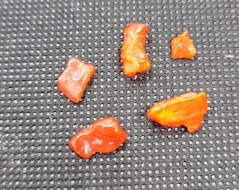 May include: Five small, irregularly shaped, polished orange gemstones are scattered on a dark, textured surface. The stones have a glossy finish, reflecting light and displaying hints of yellow and red.