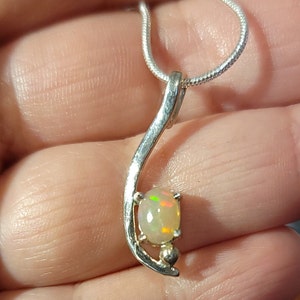 May include: A silver pendant necklace featuring an oval opal gemstone. The opal displays flashes of color and is set in a curved, silver setting. The necklace has a delicate, snake chain.